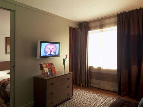 A hotel room with a television on the wall, a dresser, a window with curtains, and a bed.