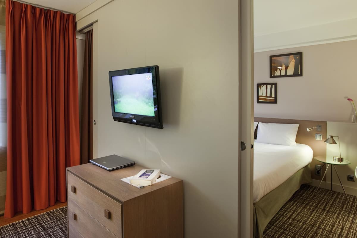 Habitación de hotel con cama, escritorio con computadora portátil, televisor montado en la pared y ventana con cortinas.