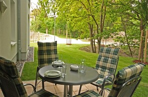 An outdoor patio with a round table, four plaid chairs, and a view of a well-maintained lawn and trees. - Haus Meeresblick FeWo Abendrot A 1.31 Ref. 132364 - FeWo Abendrot A 1.31 Ref. (Baabe)