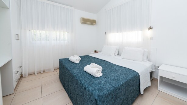 A hotel room with a bed, a nightstand, a window with sheer curtains, and an air conditioning unit. - Fig Tree Z Holiday Villas DPS09- Three Bed Bungalow (Protaras)