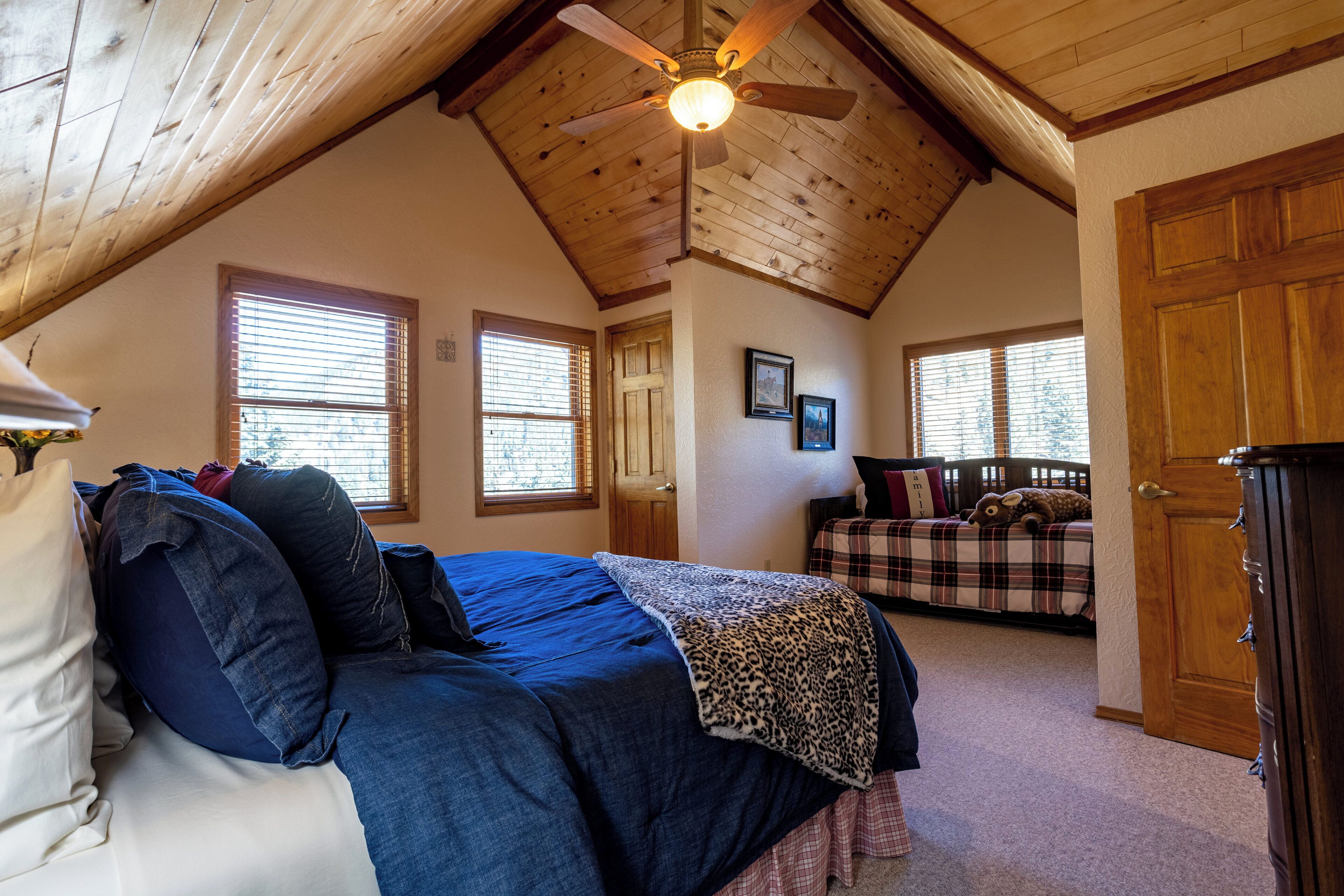 A bedroom with a sloped ceiling, a bed with a blue bedspread, a wooden dresser, and a plaid bedspread in the background.