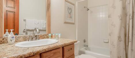 A bathroom with a granite countertop sink, wooden cabinets, a white toilet, and a bathtub with a floral shower curtain.