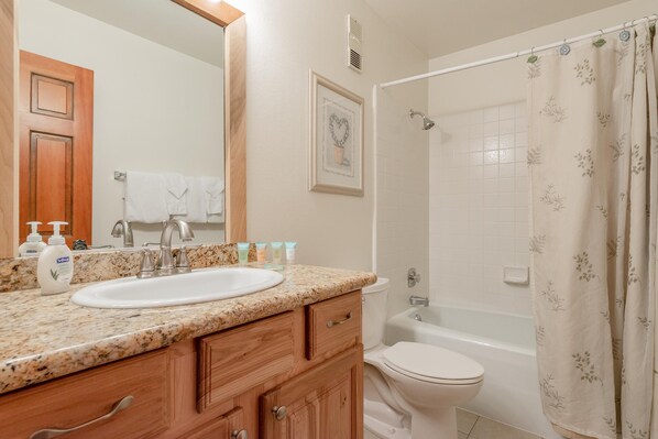Une salle de bain avec un lavabo surmonté d’un plan de travail en granit, des meubles en bois, des toilettes blanches et une baignoire agrémentée d’un rideau de douche à motifs floraux.