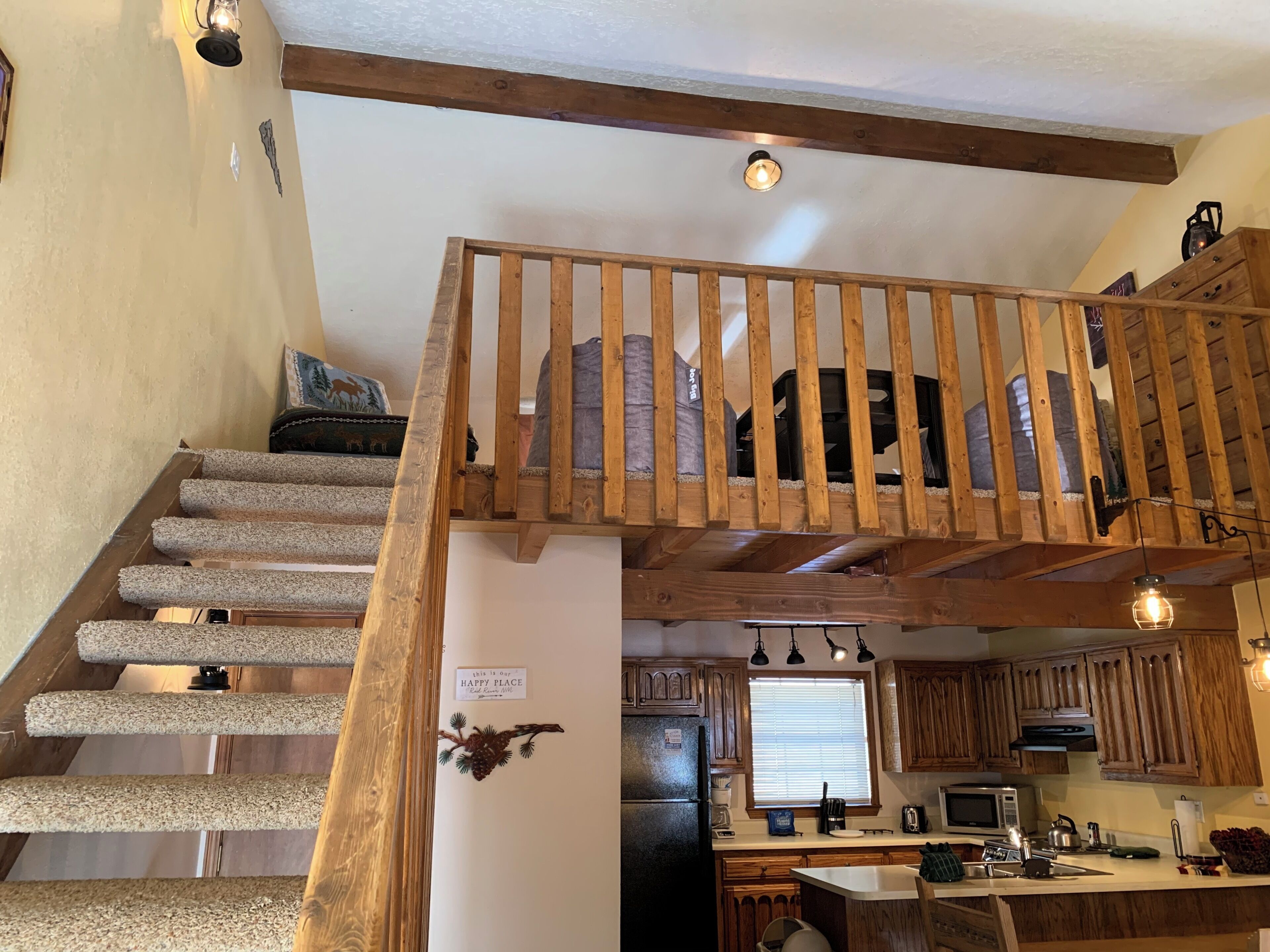 A staircase with wooden railings leading to a lofted area, a kitchen with wooden cabinets, a refrigerator, and a window with blinds.