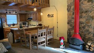 A cozy living room with a wooden dining table, chairs, a fireplace, and a kitchen area with wooden cabinets and a stone wall.