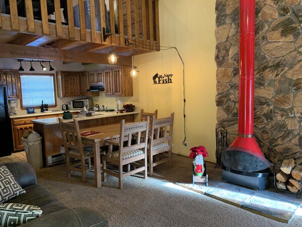 A cozy living room with a wooden dining table, chairs, a fireplace, and a kitchen area with wooden cabinets and a stone wall.