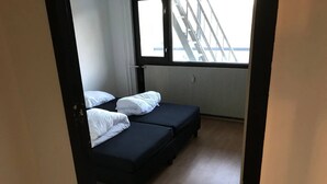 A room with two single beds, a window with a view of a building, and a radiator on the wall.