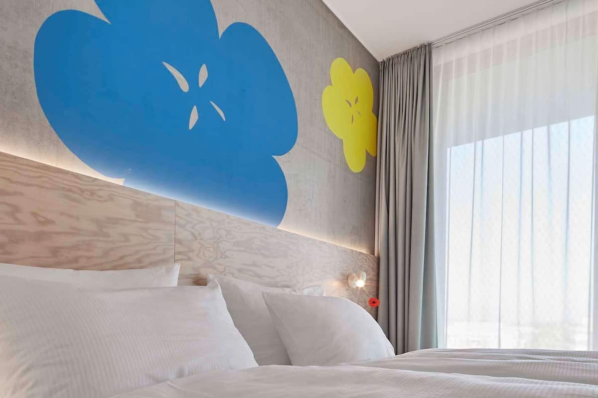 Ein Hotelzimmer mit einem großen Bett, einem Kopfteil mit floralem Muster, einem Nachttisch und einem Fenster mit Vorhängen.