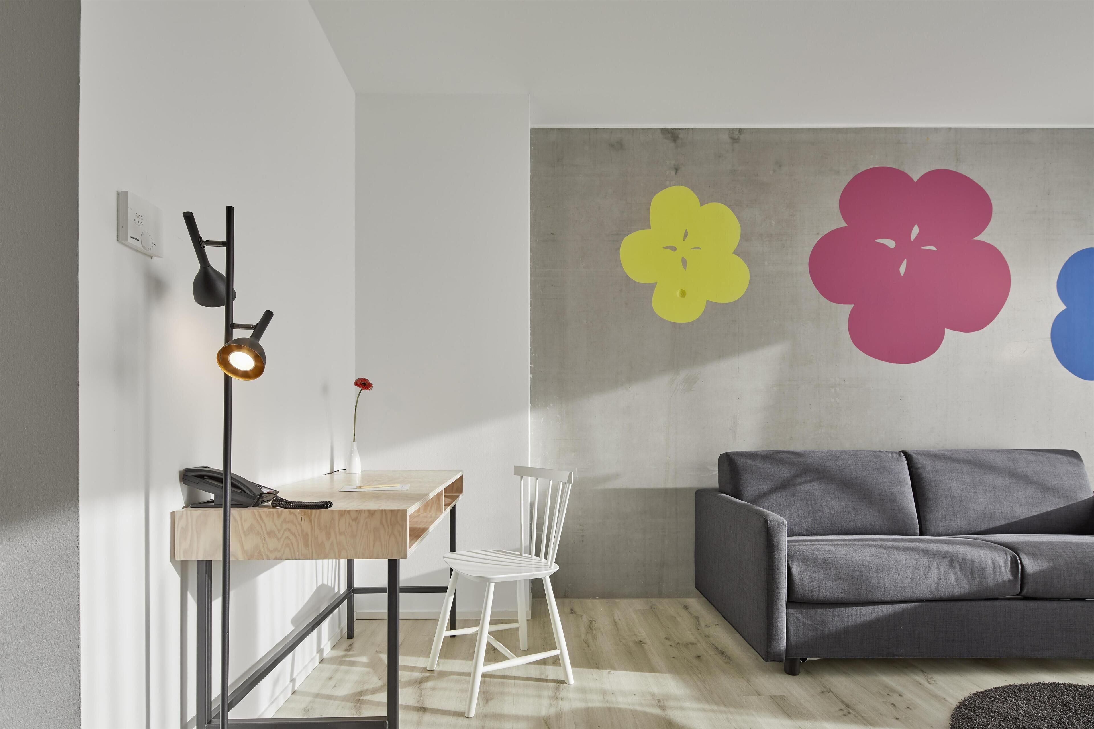 A modern living room with a grey sofa, a wooden table with a phone, a white chair, and a wall with large flower decals.