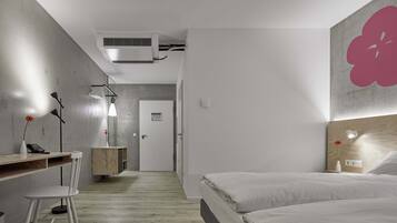 Una habitación de hotel moderna con suelo de madera, una cama con ropa blanca, un escritorio con silla, una mesita de noche con un jarrón y una obra de arte floral grande en la pared.