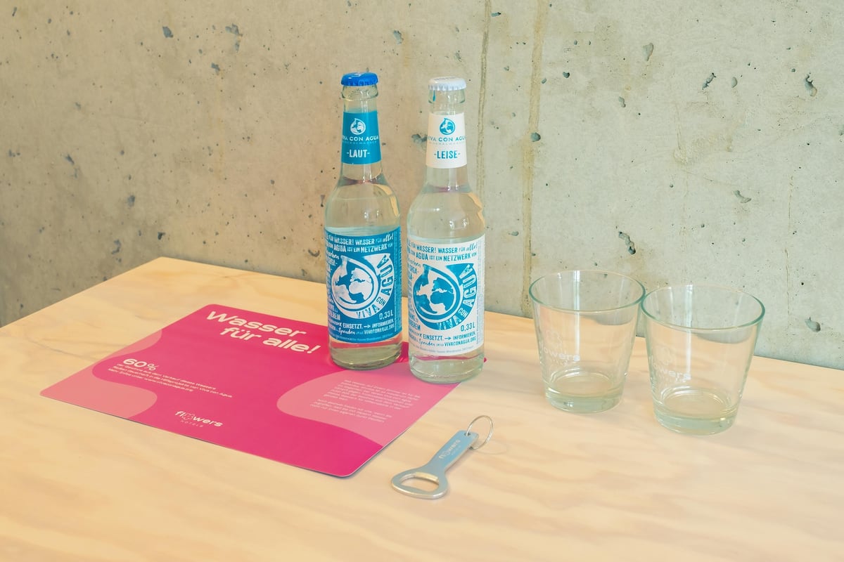 Zwei Wasserflaschen und drei Gläser auf einem Holztisch mit einer pinkfarbenen Informationstafel.