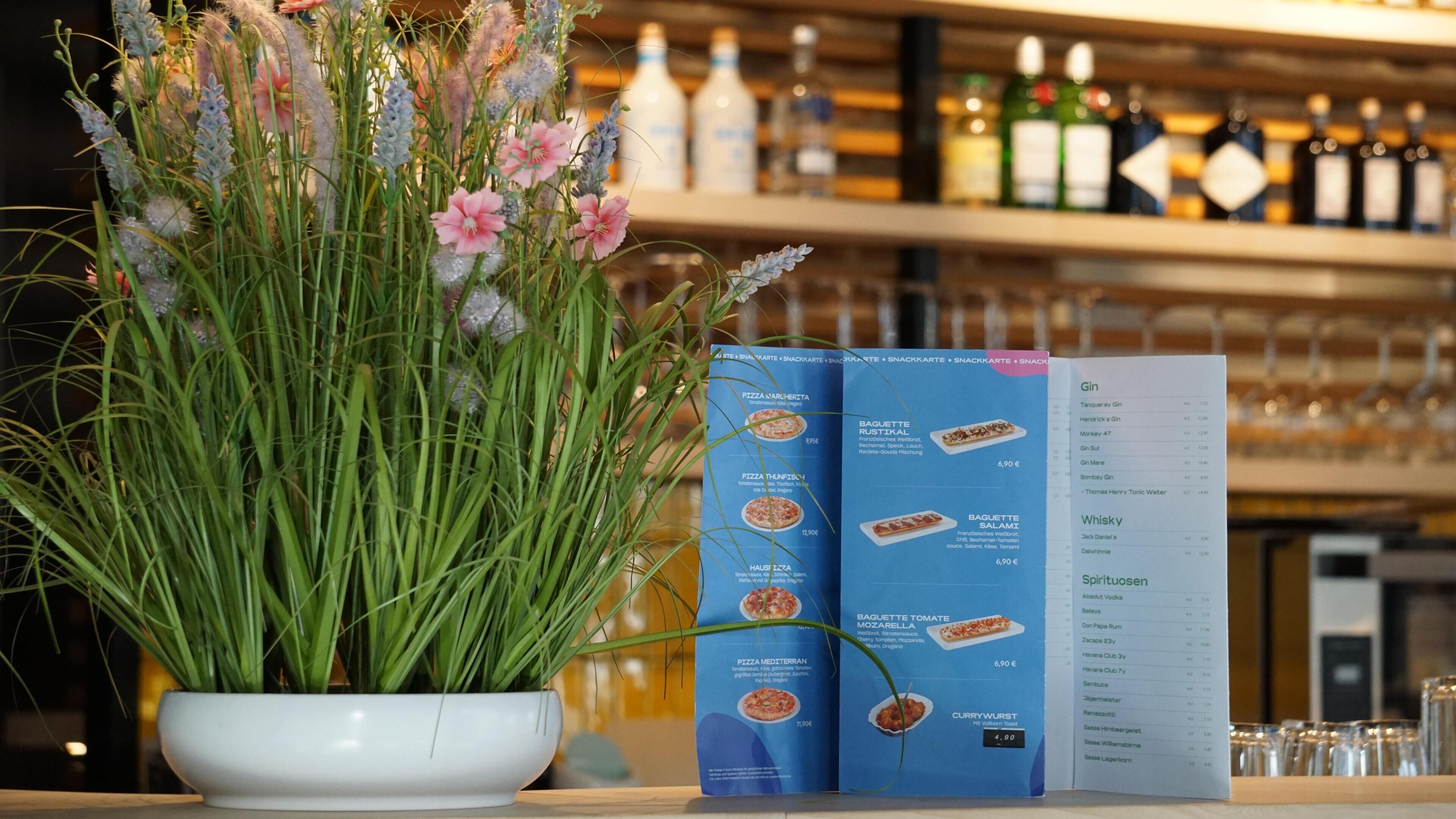 A menu with various food items and prices, a potted plant with green foliage and pink flowers, and a bar area with bottles in the background.