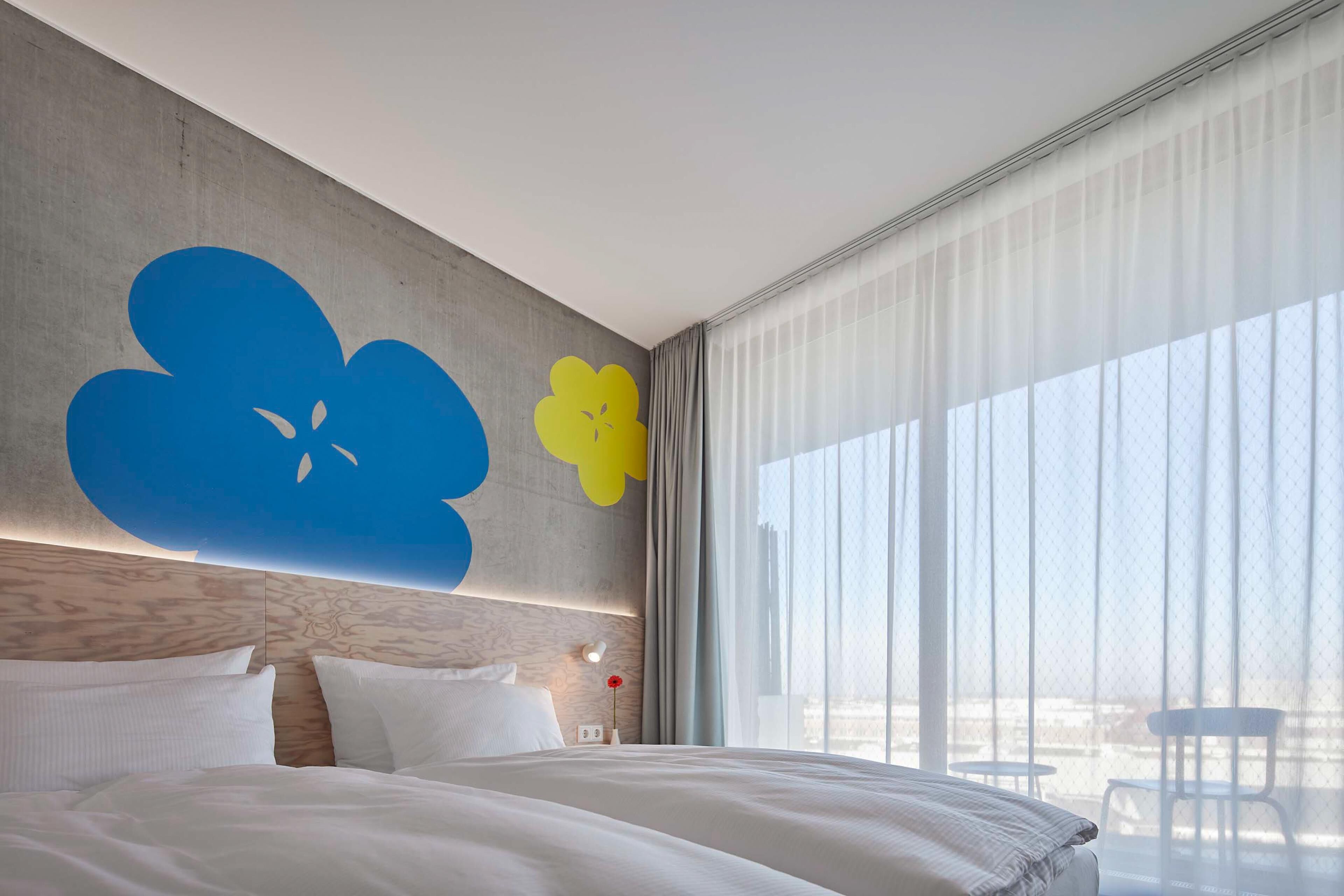 A modern hotel room with a large bed, a wooden headboard, and a large window with sheer curtains.