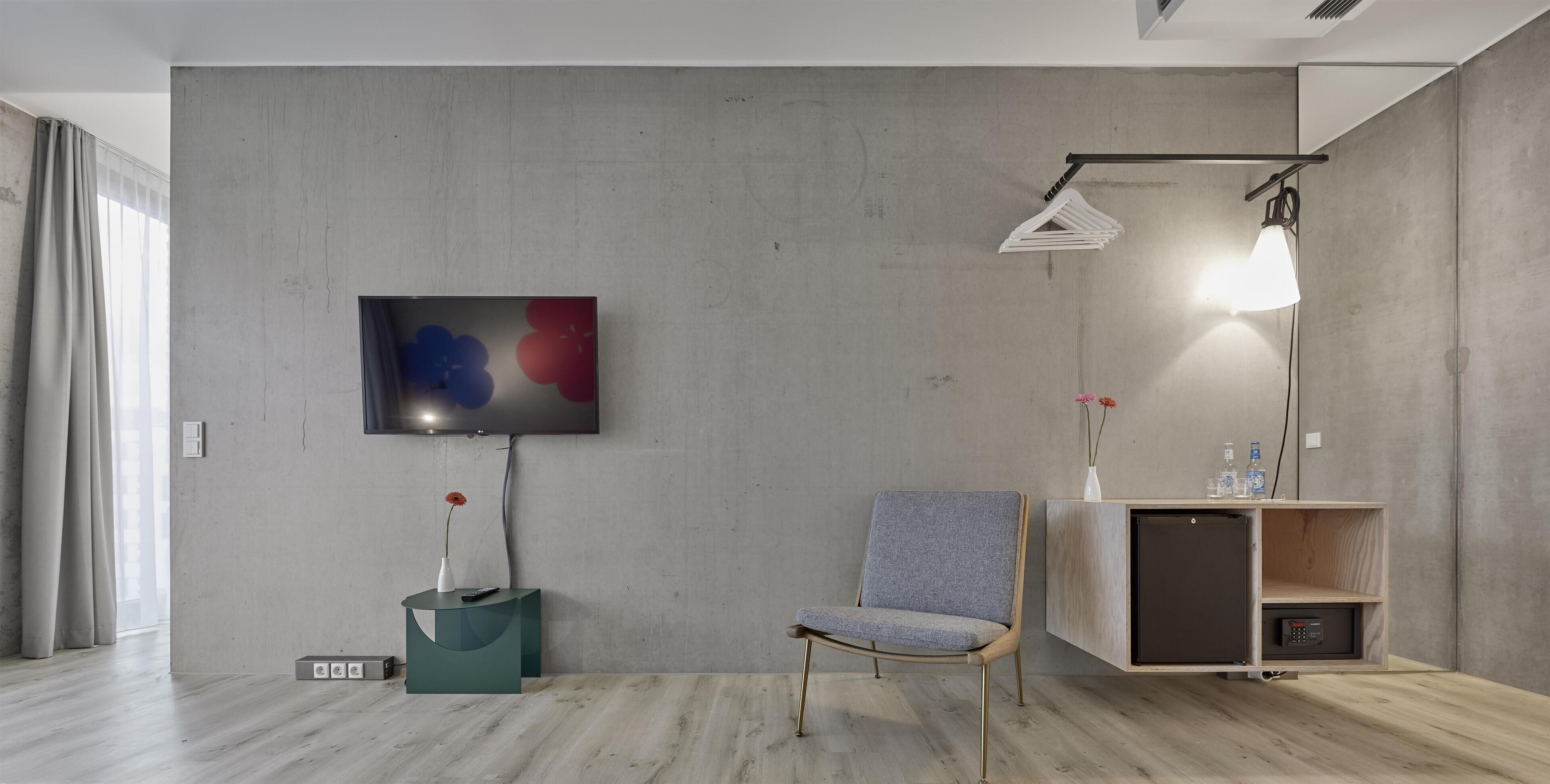 Ein modernes Hotelzimmer mit grauer Betonwand, einem Flachbildfernseher, einem grau gepolsterten Sessel, einem Holznachttisch mit Vase und Flaschen sowie einer Stehlampe.