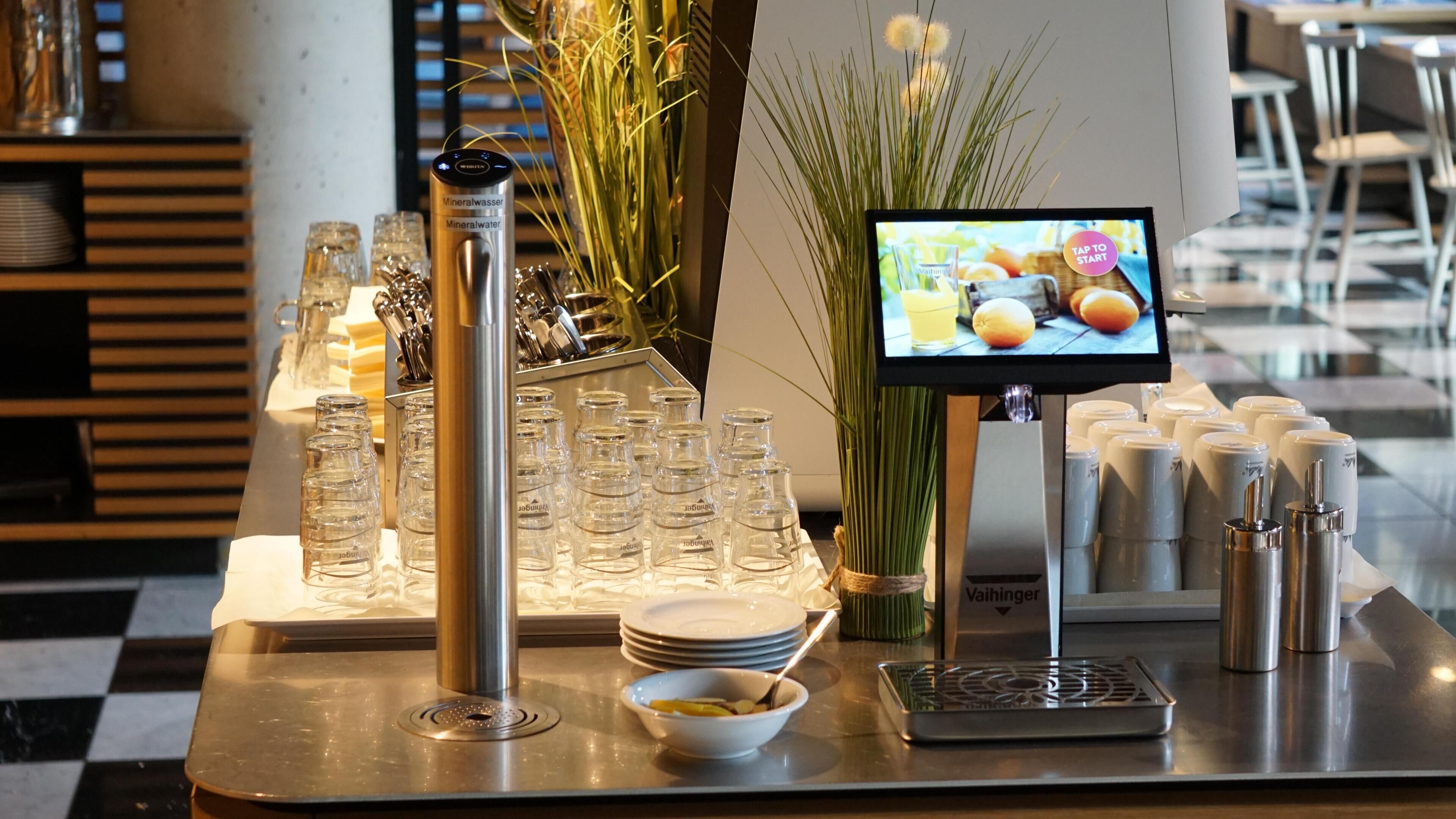 Montaje tipo bufé con cristalería transparente, dispensador de agua mineral, pantalla digital con el menú y una máquina de café Vaishinger.