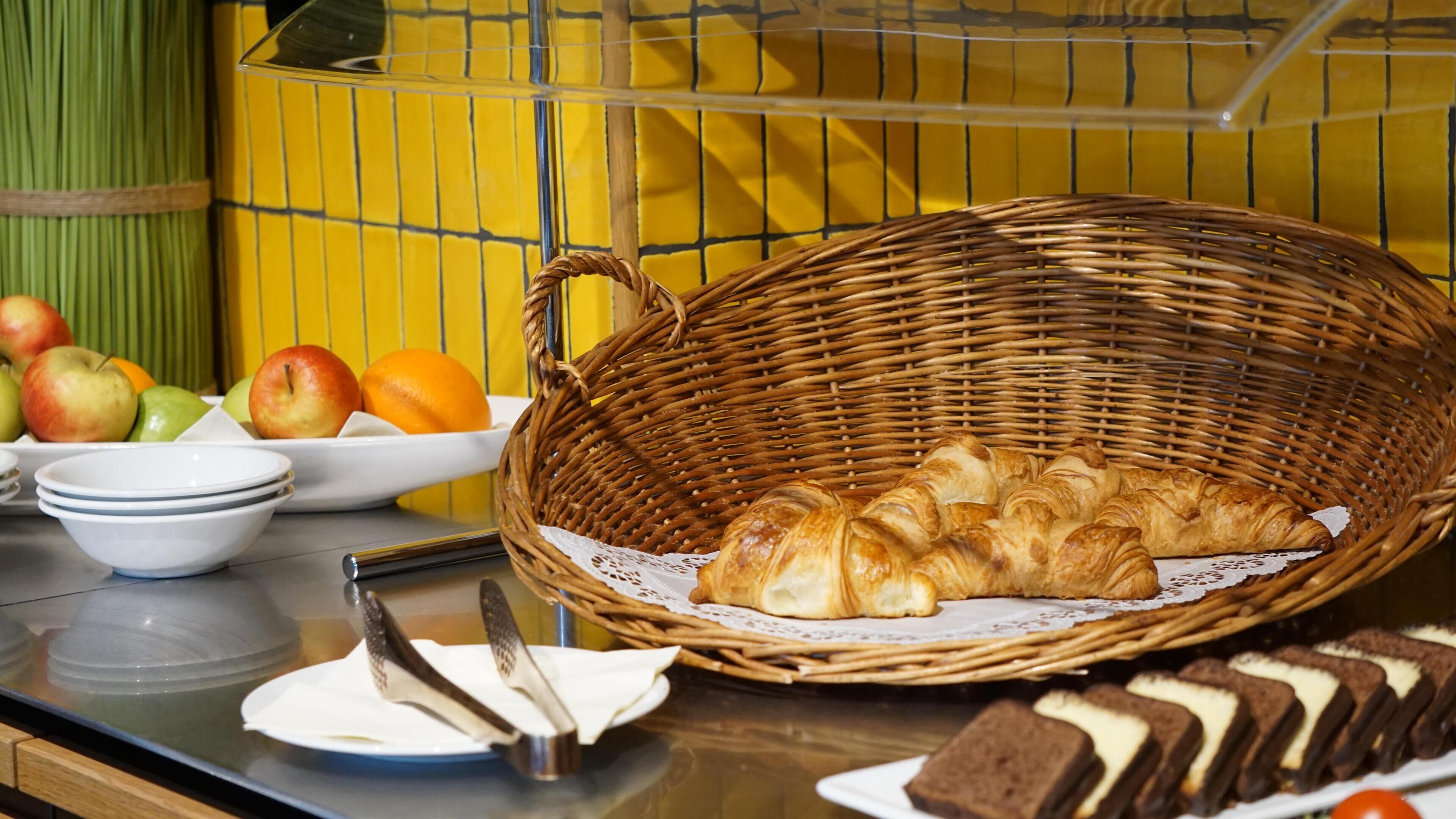 Ein Frühstücksbuffet mit einem Korbwannen-Arrangement, das Croissants, einen Teller Äpfel und einen Teller Orangen enthält.