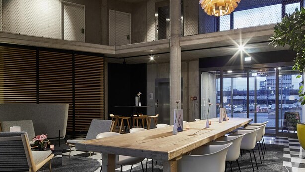 A modern office space with a large wooden table, white chairs, and hanging pendant lights. - Flowers Hotel Essen (Essen)