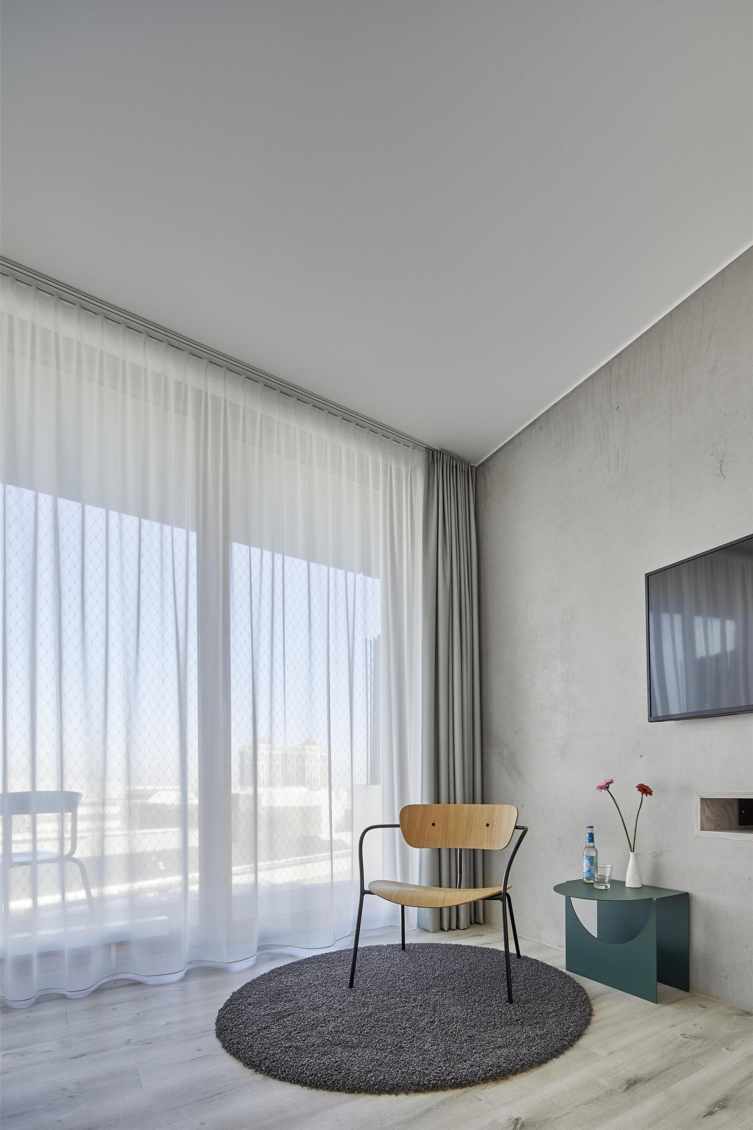 A modern room with a round rug, a wooden chair, a side table with a vase and flowers, and a large window with sheer curtains.