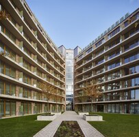 Un moderno edificio de varios pisos con un patio central, que cuenta con balcones con plantas verdes y un camino que conduce a la entrada.
