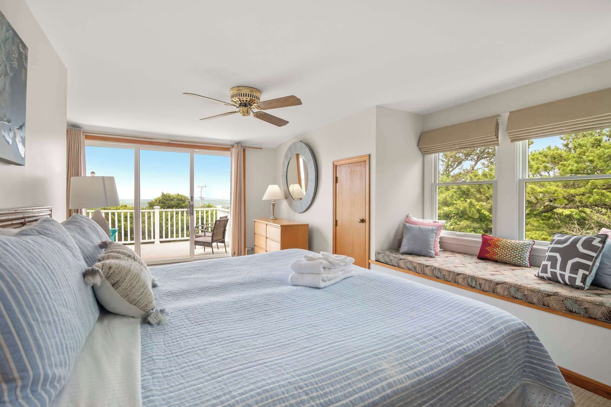 A bedroom with a large bed, a ceiling fan, a window seat with cushions, and a view of the outdoors.