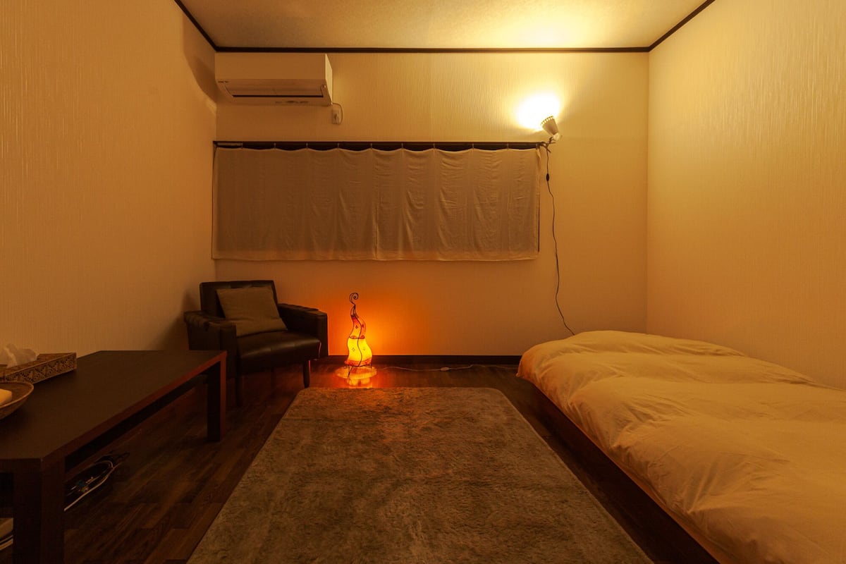 Une petite pièce faiblement éclairée, avec un lit simple, une chaise, une table et un climatiseur fixé au mur.
