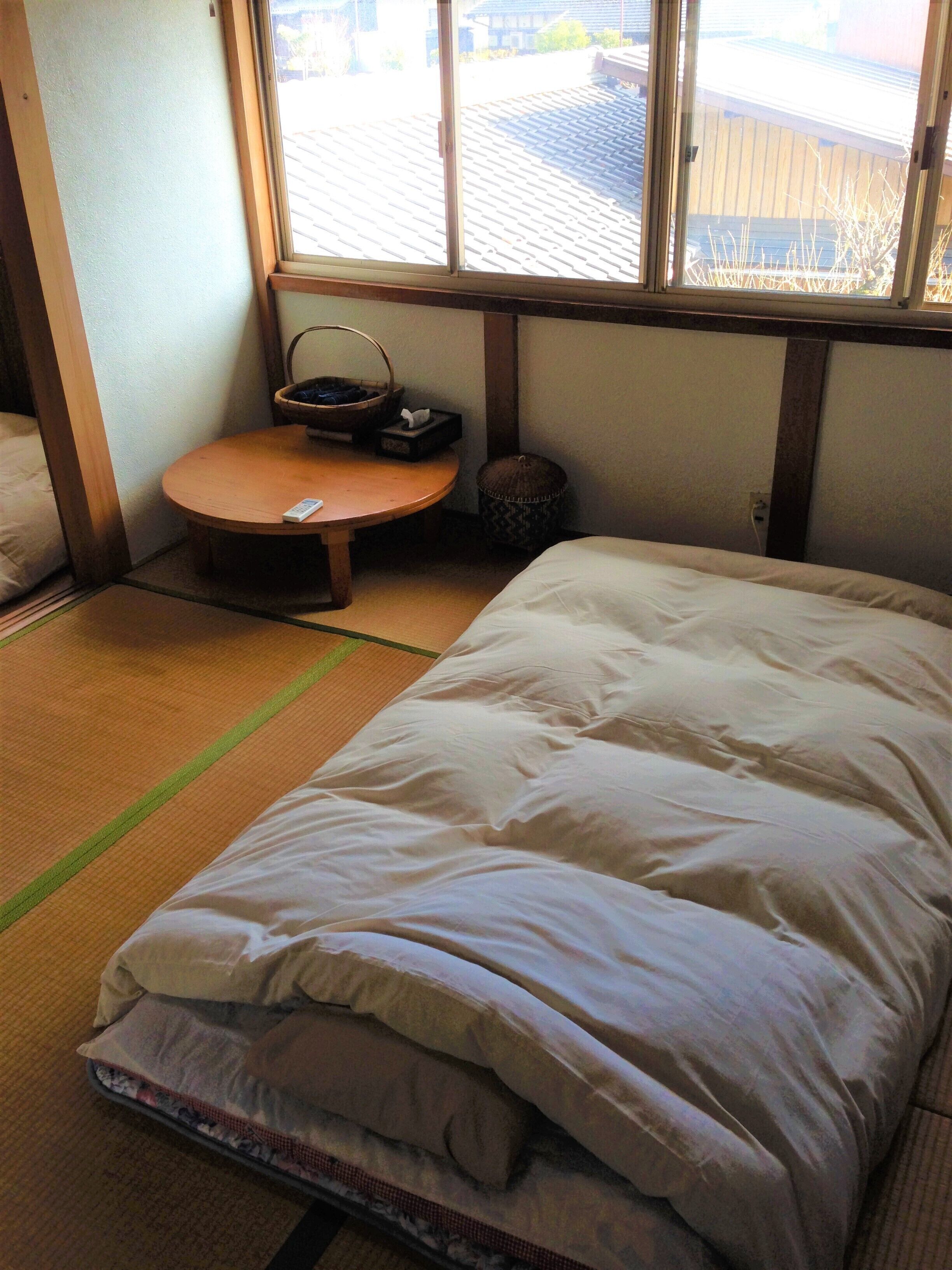 A room with a bed, a small round table with a basket and tissue box, and a window with a view of a building and trees.