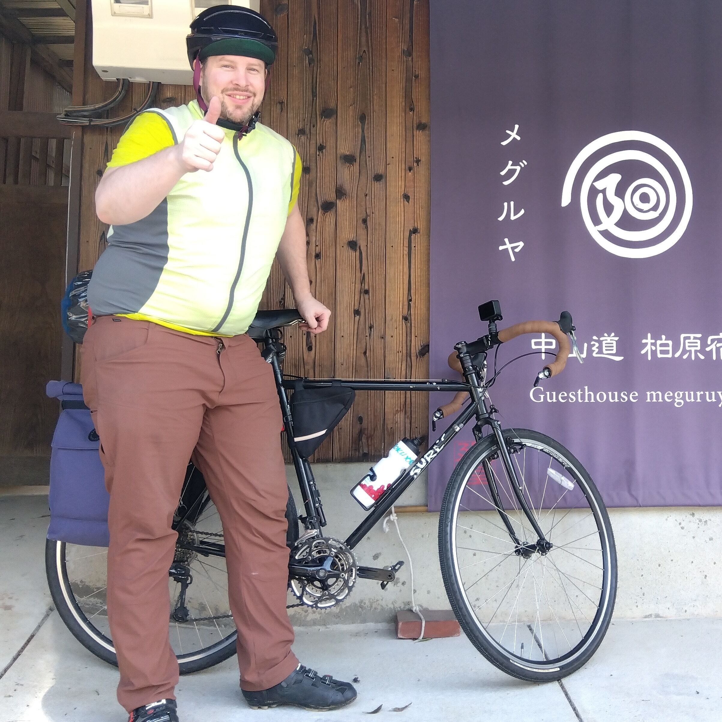 A person standing next to a bicycle with a water bottle attached to the frame. There is a purple banner with Japanese text and a logo in the background.