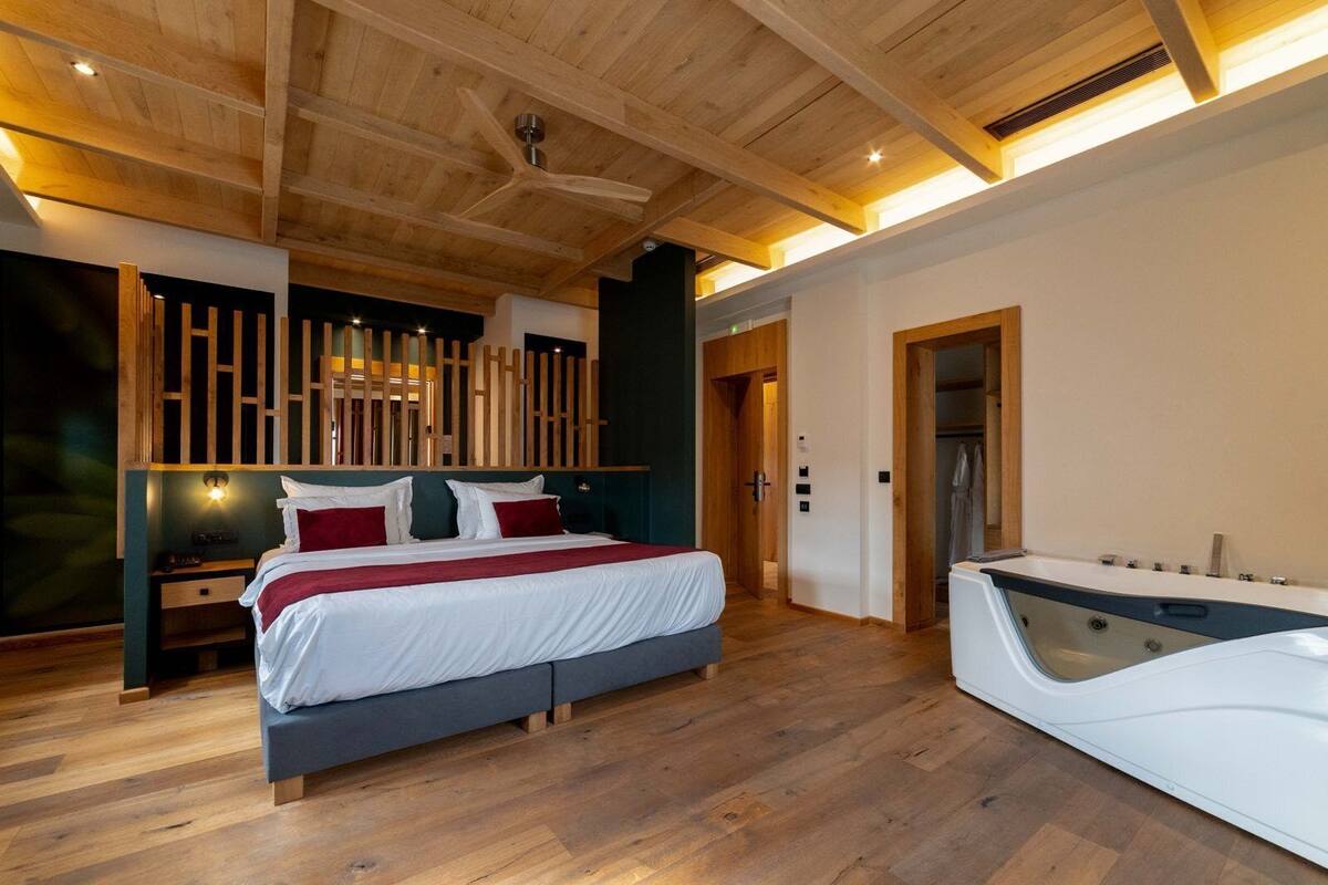 A modern bedroom with a large bed, wooden ceiling beams, and a built-in jacuzzi.