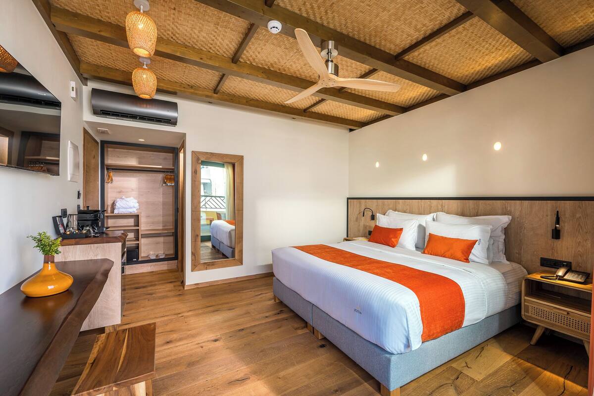 A modern hotel room with a large bed, wooden flooring, a desk with a plant, a ceiling fan, and a view of the outside.