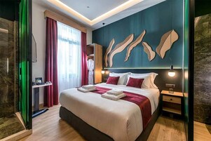 Quarto de hotel moderno com uma cama grande, uma mesa de cabeceira de madeira, uma luminária fixada na parede e uma janela com cortinas.