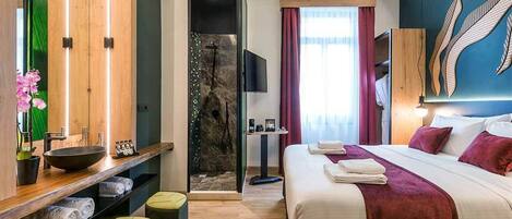 Una habitación de hotel moderna con una cama grande, una tocador de madera con lavamanos, zona de ducha y televisión.