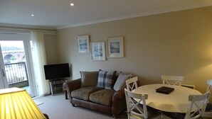 A living room with a sofa, a TV, a dining table with chairs, and framed pictures on the wall. - Tay Pearls - Tay Chambers Apartment (Perth)