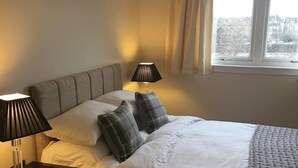 A neatly made bed with white linens and a gray headboard, flanked by two bedside tables with lamps, a window with curtains, and a view of a snowy landscape. - Tay Pearls - Tay Chambers Apartment (Perth)