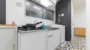 Uma cozinha compacta com fogão a gás, pia e lava-louças. Há uma janela acima da pia e uma porta que dá para outro cômodo. O chão tem um design de azulejo com padrão geom étrico.