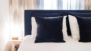 Een bed met een donker hoofdbord en twee kussens, één donker en één wit, in een kamer met een lichtgekleurde muur en een raam met gordijnen.