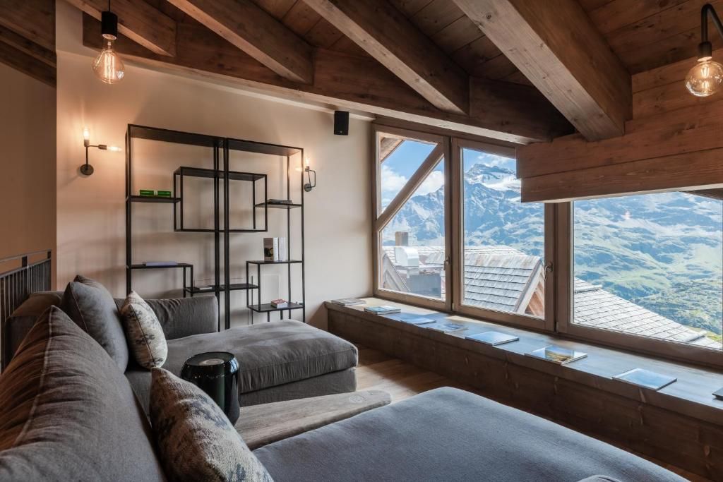 Ein gemütliches Zimmer mit Holzboden, einer Couch, einem kleinen Tisch und einem Bücherregal. Ein großes Fenster bietet Blick auf Berge.