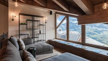 Ein gemütliches Zimmer mit Holzboden, einer Couch, einem kleinen Tisch und einem Bücherregal. Ein großes Fenster bietet Blick auf Berge.