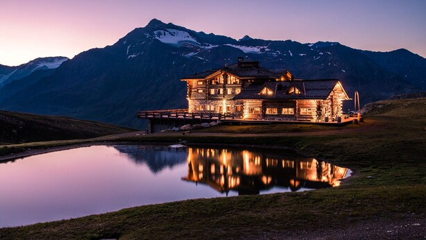 Um hotel em estilo cabana de tronco com reflexo em um lago, rodeado por montanhas.