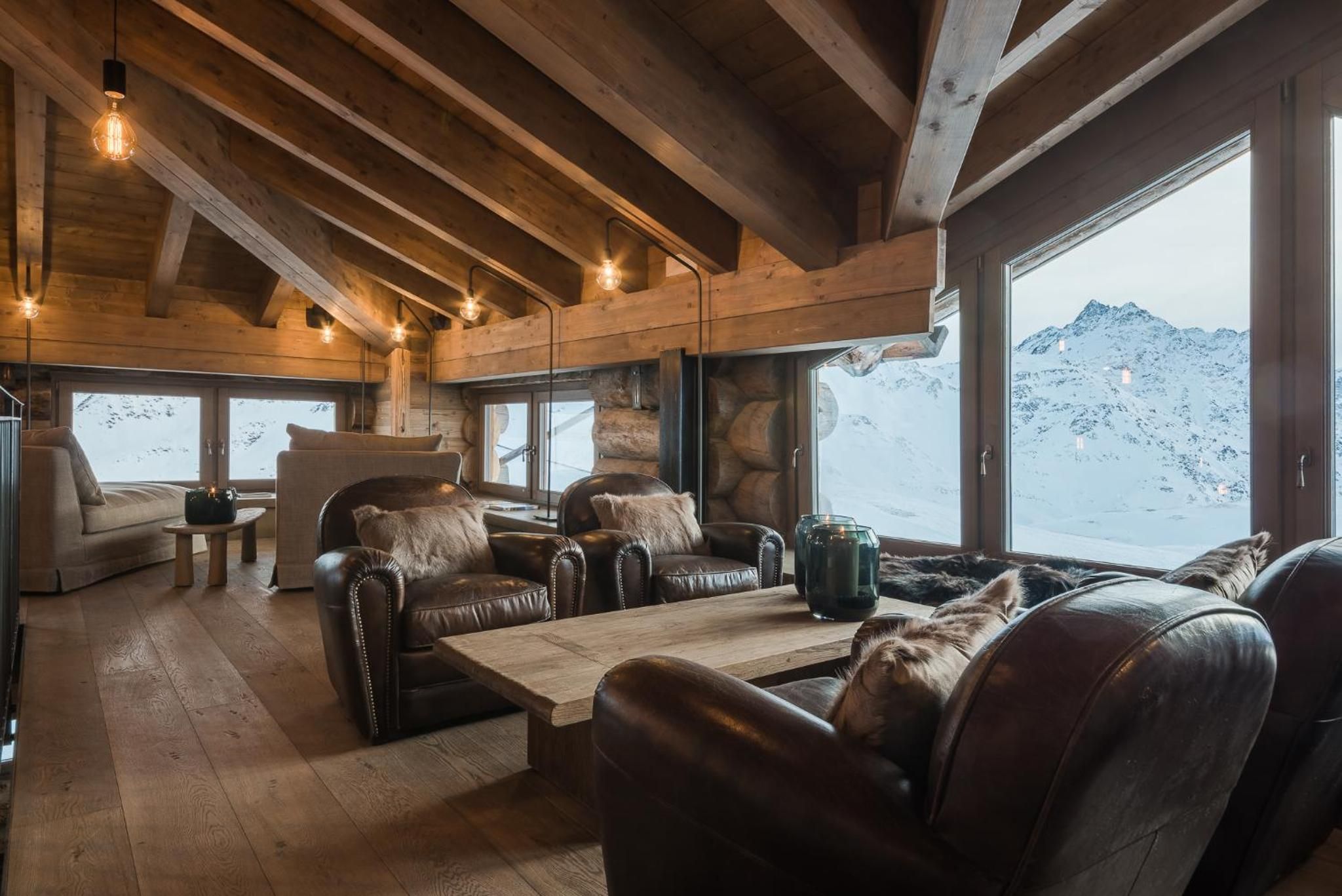Interior acogedor de una cabaña con vigas de madera, sofás de cuero, una mesa de centro de madera y ventanas amplias con vistas a la montaña.