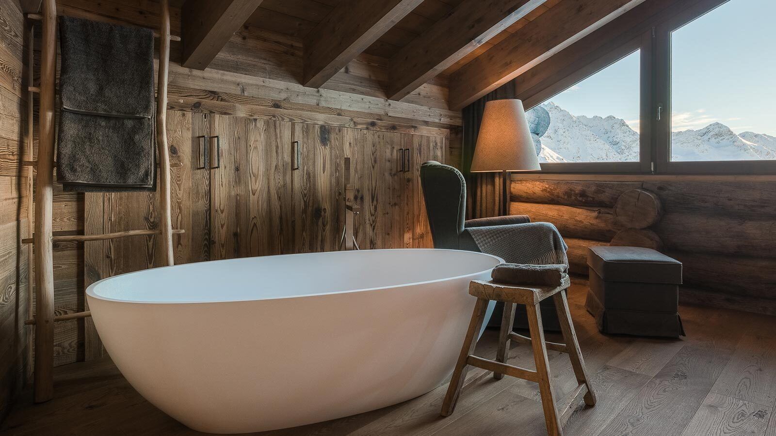 Une salle de bain rustique, aménagée dans une cabane en rondins, avec une baignoire sur pieds, des murs en bois et une vue sur des montagnes enneigées.