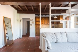 A room with a white sofa, a wooden ceiling, and a kitchen area visible through an open door.