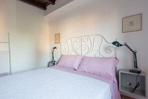 A bed with a white headboard, a pink bedspread, and two pillows. There is a nightstand with a lamp and a small shelf with a power outlet.