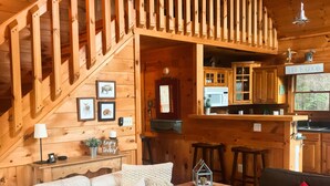 Een houten cabin interieur met een trap, een woonkamer met een witte en een donkere bank, een houten eettafel en een keuken met houten kasten en een raam.