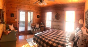 Une chambre confortable dans un chalet en bois, avec une couverture à carreaux sur le lit, une table de chevet avec une lampe, une chaise avec des coussins et une petite table avec des livres.