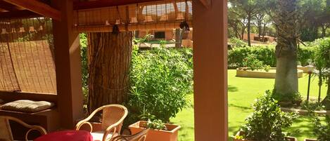 A covered patio with wicker furniture and a red tablecloth, overlooking a well-maintained garden with various plants and a lawn.