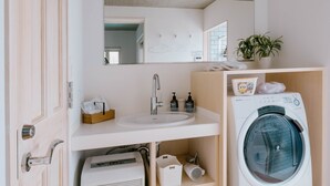 A compact, modern bathroom with a built-in sink, a mirror, a washing machine, and a small shelf with toiletries. - Casablanca Pool House (Kamakura)