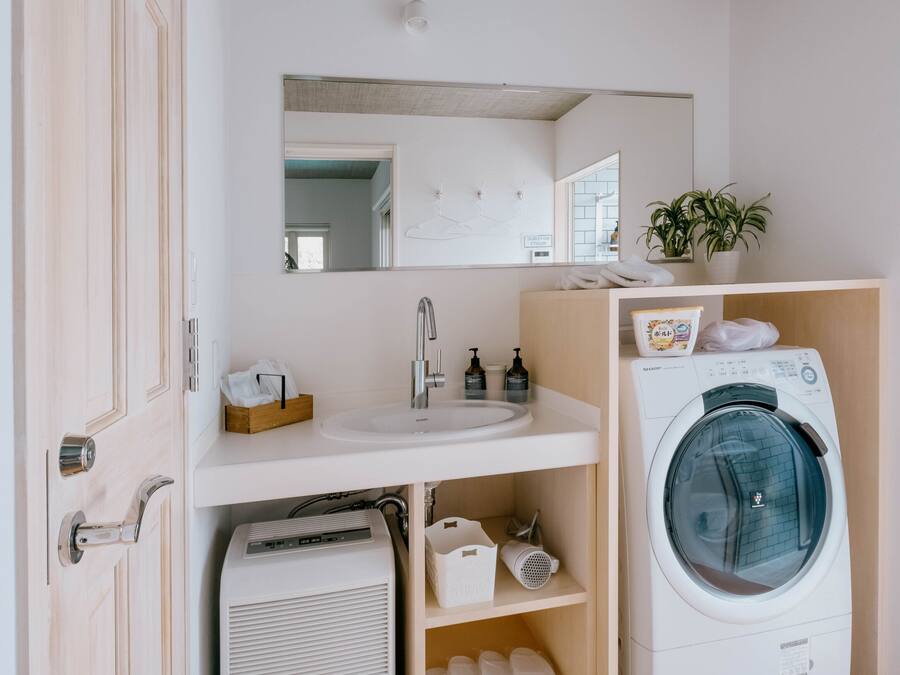 A compact, modern bathroom with a built-in sink, a mirror, a washing machine, and a small shelf with toiletries.