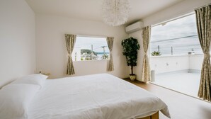 A bedroom with a large bed, a potted plant, a window with curtains, and a view of the outside. - Casablanca Pool House (Kamakura)
