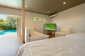 Quarto de hotel moderno com uma cama grande, duas poltronas brancas, uma televisão e vista para uma piscina.