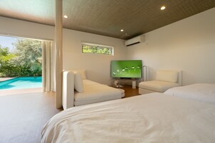 A modern hotel room with a large bed, two white armchairs, a television, and a view of a swimming pool.